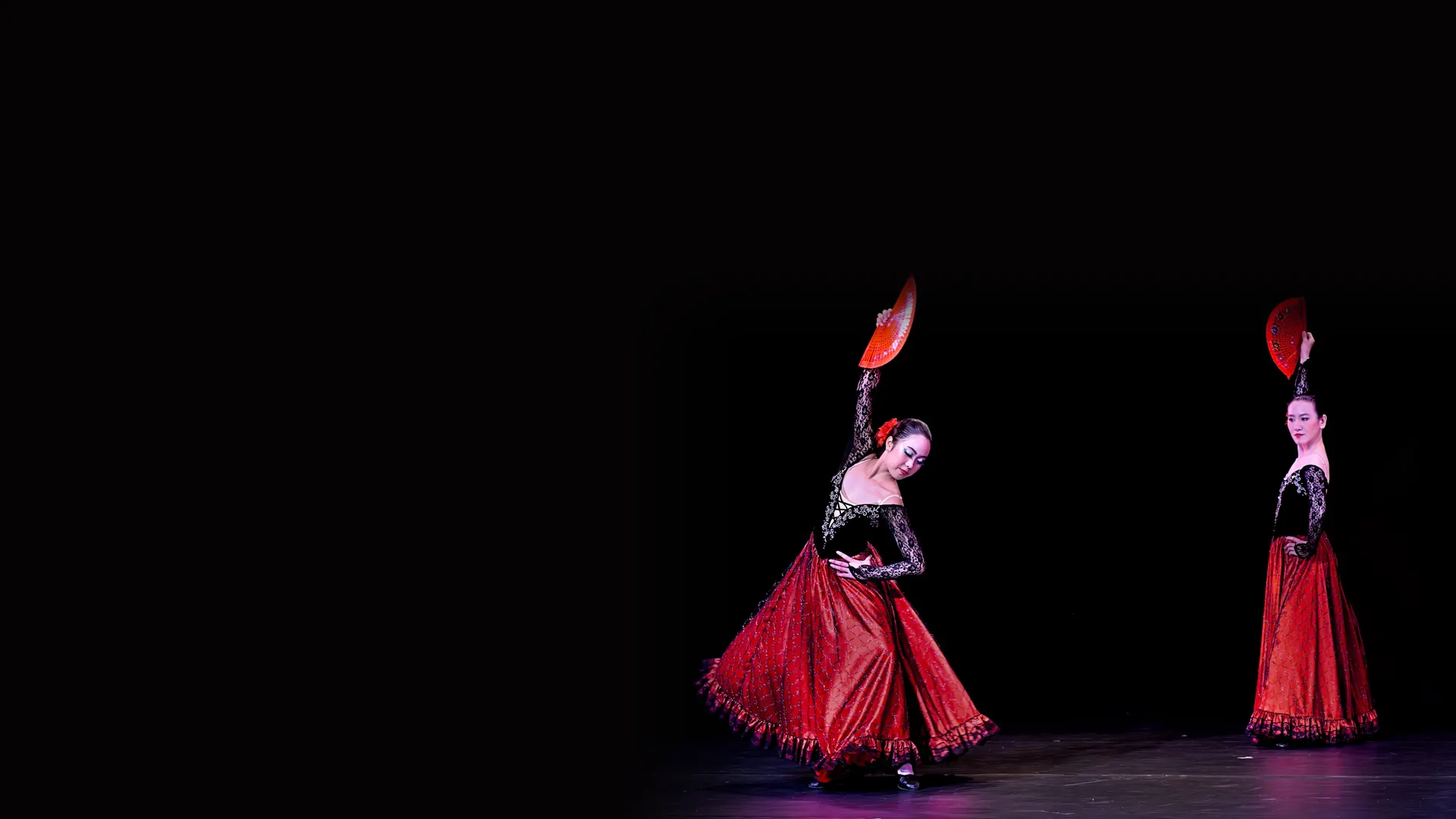 Dvě flamencové tanečnice v červených volánových sukních a černých krajkových topů, každá s červeným vějířem, na tmavém jevišti.